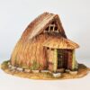 A 28mm scale 3D printed model of a Koyane-Minka, a small, circular Japanese folk house. The model features a rounded, bell-shaped thatched roof that curves toward the ground, a small sheltered entrance supported by two timber pillars, and a stone-lined foundation. The design reflects a primitive, organic mountain hermitage or rural storage hut.