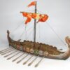A fully painted 3D printed model of a Viking Drakkar longship. The model features a detailed wooden texture, a dragon head prow, a row of colorful shields along the gunwale, oars, and a single mast with a furled orange sail.