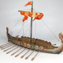 A fully painted 3D printed model of a Viking Drakkar longship. The model features a detailed wooden texture, a dragon head prow, a row of colorful shields along the gunwale, oars, and a single mast with a furled orange sail.