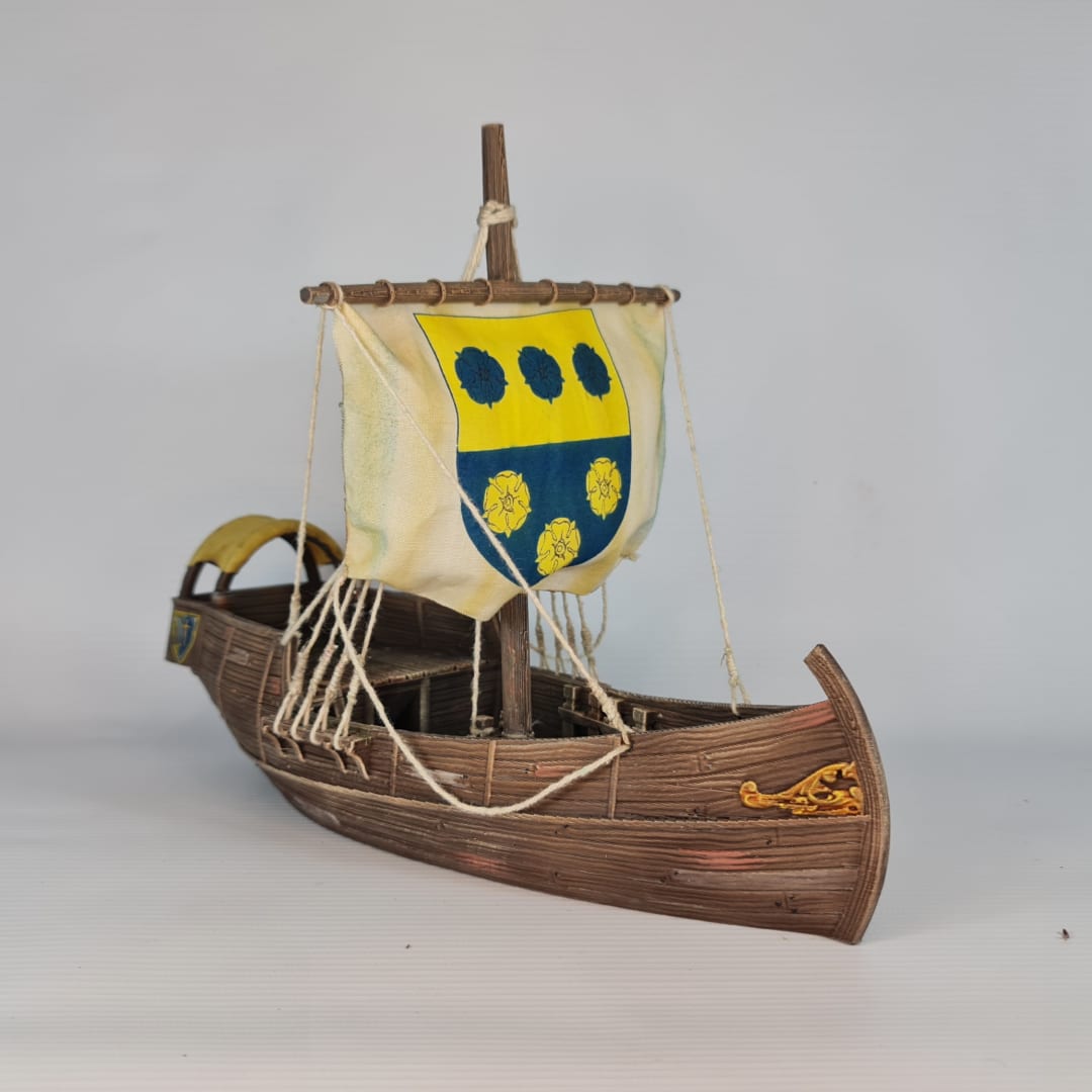 3D printed and painted model of a medieval merchant cog ship, front view. The ship has a detailed wooden hull, a single mast, and a square sail with a blue and yellow coat of arms.