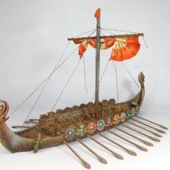 A fully painted 3D printed model of a Viking drakkar longship, featuring a carved dragon head prow, a line of colourful shields along the side, numerous oars, and a furled orange and white sail on a central mast.