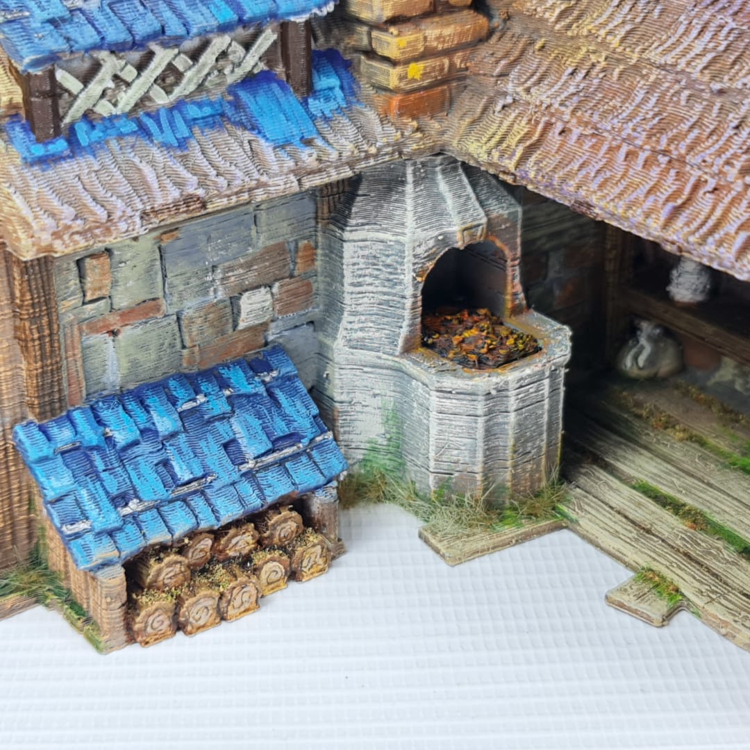 3D printed and painted miniature of a medieval blacksmith building with a stone foundation, wooden wattle-and-daub walls, a side waterwheel, thatched awnings, and a striking blue tiled roof.