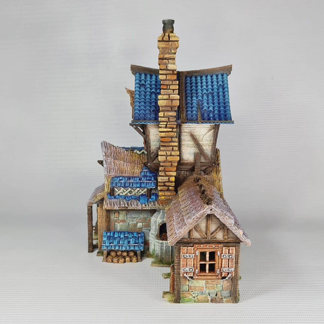 3D printed and painted miniature of a medieval blacksmith building with a stone foundation, wooden wattle-and-daub walls, a side waterwheel, thatched awnings, and a striking blue tiled roof.