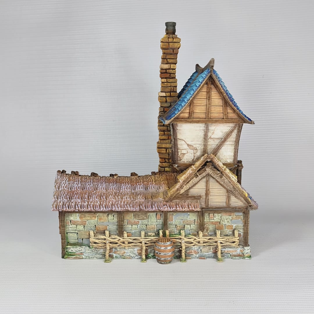 3D printed and painted miniature of a medieval blacksmith building with a stone foundation, wooden wattle-and-daub walls, a side waterwheel, thatched awnings, and a striking blue tiled roof.