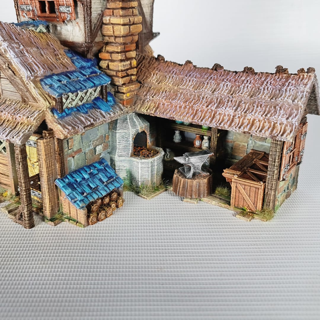 3D printed and painted miniature of a medieval blacksmith building with a stone foundation, wooden wattle-and-daub walls, a side waterwheel, thatched awnings, and a striking blue tiled roof.