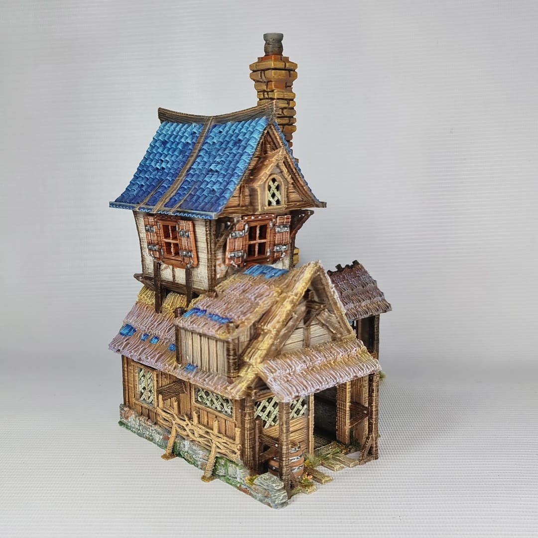 3D printed and painted miniature of a medieval blacksmith building with a stone foundation, wooden wattle-and-daub walls, a side waterwheel, thatched awnings, and a striking blue tiled roof.