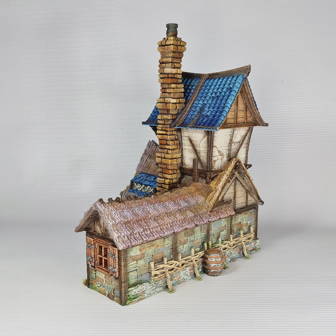 3D printed and painted miniature of a medieval blacksmith building with a stone foundation, wooden wattle-and-daub walls, a side waterwheel, thatched awnings, and a striking blue tiled roof.