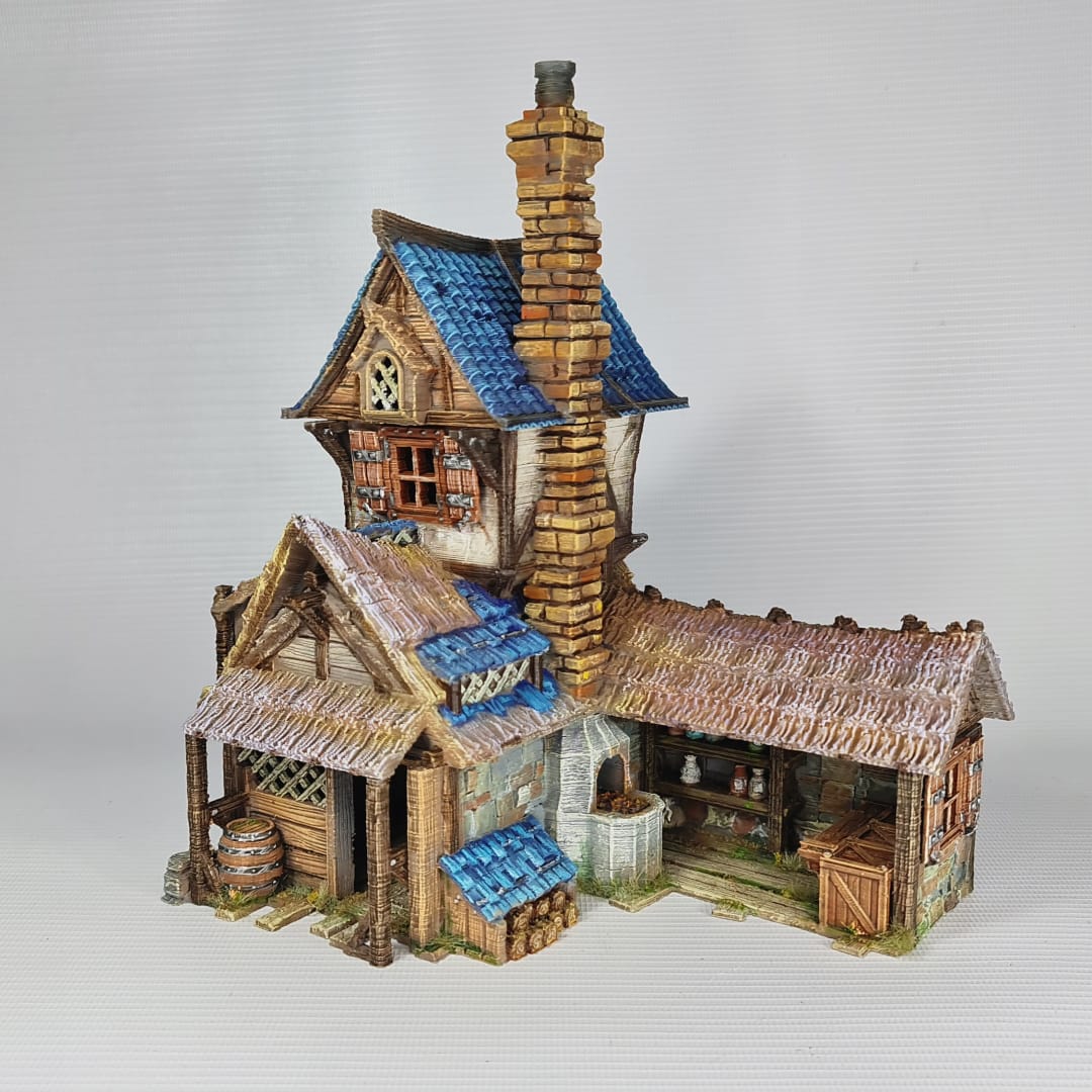 3D printed and painted miniature of a medieval blacksmith building with a stone foundation, wooden wattle-and-daub walls, a side waterwheel, thatched awnings, and a striking blue tiled roof.