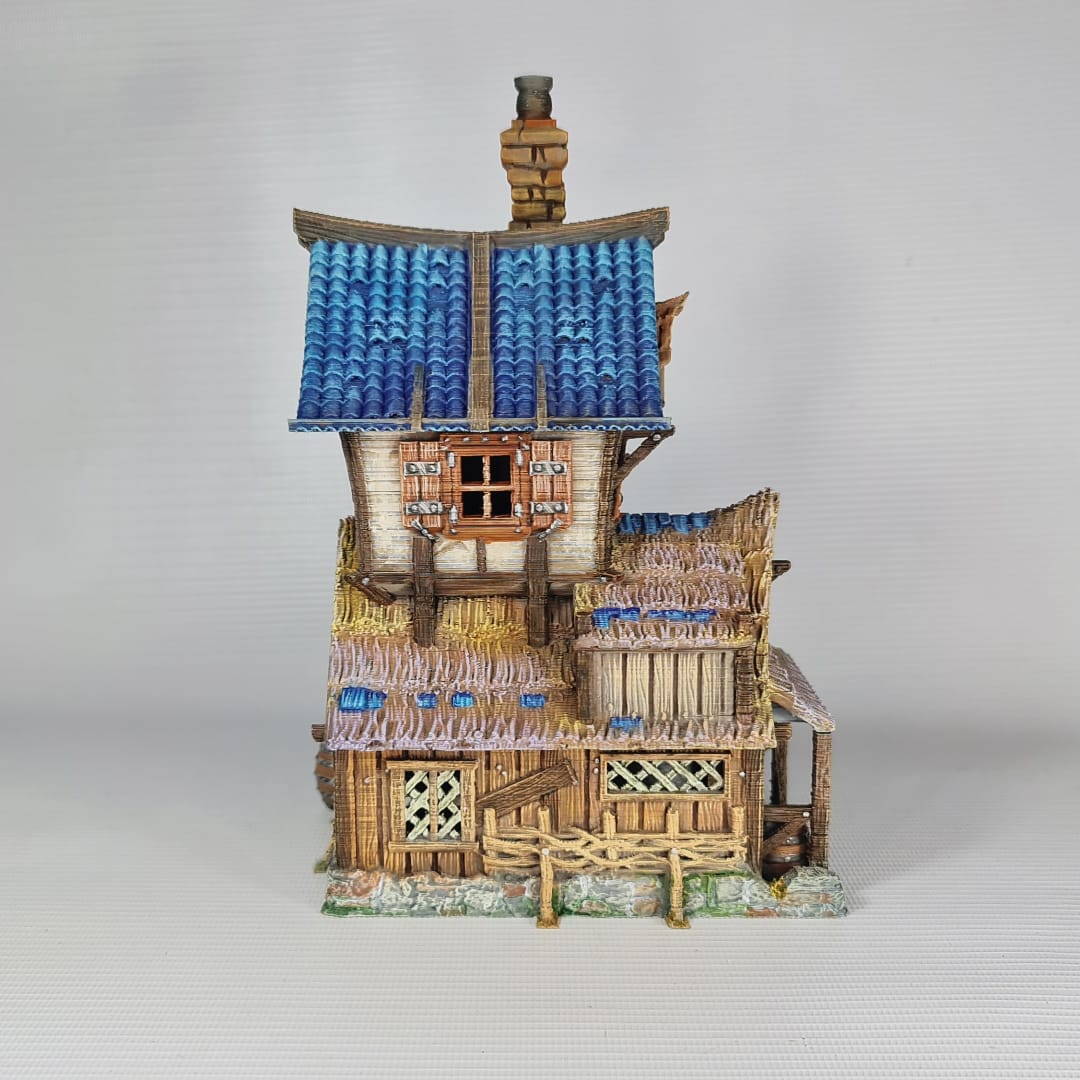 3D printed and painted miniature of a medieval blacksmith building with a stone foundation, wooden wattle-and-daub walls, a side waterwheel, thatched awnings, and a striking blue tiled roof.