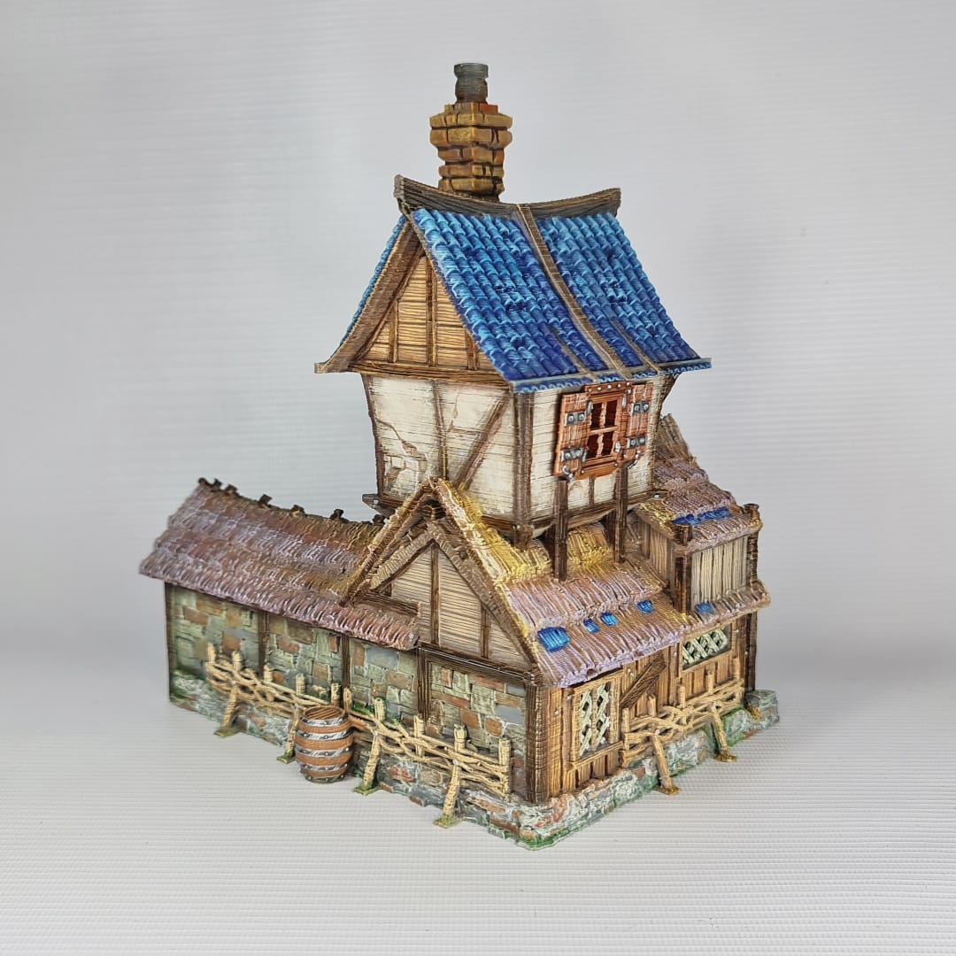 3D printed and painted miniature of a medieval blacksmith building with a stone foundation, wooden wattle-and-daub walls, a side waterwheel, thatched awnings, and a striking blue tiled roof.